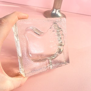 Puede incluir: Un cenicero de vidrio transparente con un diseño de pájaro en relieve. El cenicero es cuadrado y tiene una superficie texturizada.