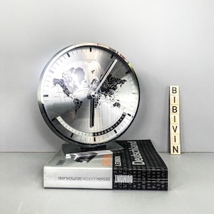 May include: A silver world clock with a black map of the world and black hour and minute hands. The clock is sitting on a black book with the text "DESIGNLEXIKON DEUTSCHLAND DUMONT LEMON DESIGN" on the spine.