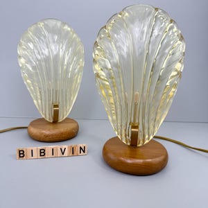 Könnte beinhalten: Zwei Vintage-Lampen in Muschelform mit warmgelbem Schein. Jede Lampe hat einen transparenten, strukturierten Glasschirm und einen Holzsockel. Die Lampen sind durch eine goldfarbene Metallhalterung und ein braunes Kabel verbunden. Das Wort "BIBIVIN" ist mit Holzbuchstaben geschrieben.