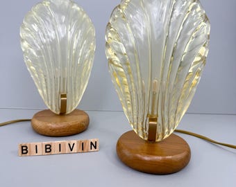 May include: Two vintage shell-shaped lamps with a warm yellow glow. Each lamp features a clear, textured glass shade and a wooden base. The lamps are connected by a gold-colored metal support and a brown cord. The word "BIBIVIN" is spelled out in wooden letter tiles.