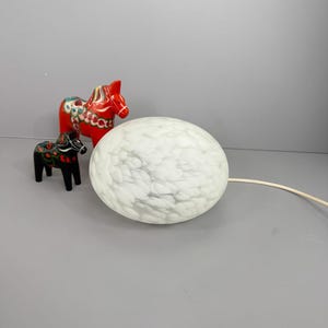 May include: A white, spherical lamp with a marbled design, accompanied by two wooden Dala horses. One horse is red with green and white details, and the other is black with floral accents. The lamp has a white cable.