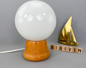 Puede incluir: Una lámpara de mesa blanca en forma de globo con base de madera. La lámpara se combina con una escultura de velero dorada. La palabra "BIBIVIN" está escrita con letras de madera.