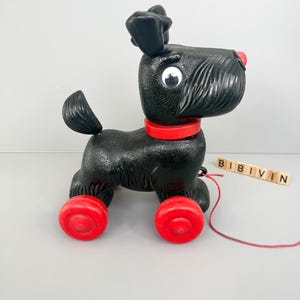 Può includere: Un cane giocattolo nero da tirare con ruote rosse, un collare rosso e un naso rosso. Il cane ha un corpo strutturato e una coda. La parola "BIBIVIN" è scritta con lettere di legno.