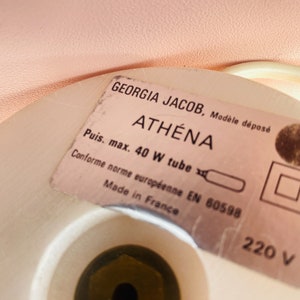 May include: Close-up of a white lamp base with a label that reads "GEORGIA JACOB, Mod&egrave;le d&eacute;pos&eacute; ATH&Eacute;NA Puis. max. 40 W tube Conforme norme europ&eacute;enne EN 60598 Made in France 220 V".