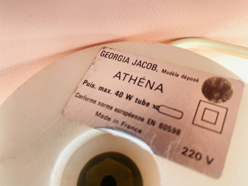 May include: Close-up of a white lamp base with a label that reads "GEORGIA JACOB, Mod&egrave;le d&eacute;pos&eacute; ATH&Eacute;NA Puis. max. 40 W tube Conforme norme europ&eacute;enne EN 60598 Made in France 220 V".