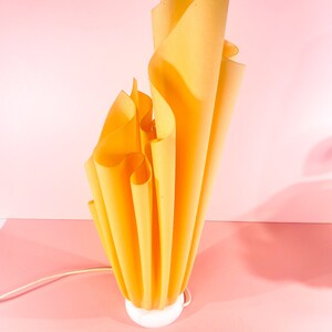 May include: A yellow, pleated lamp with a white base. The lamp is shaped like a flame and is lit up.