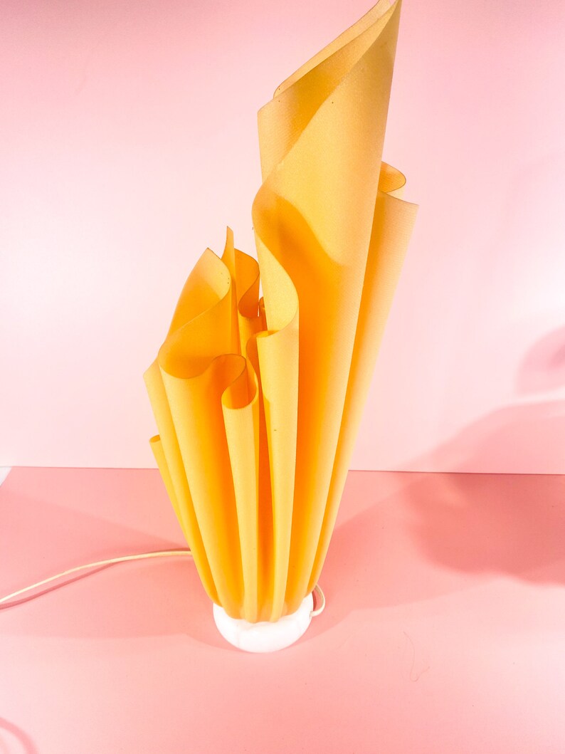 May include: A yellow, pleated lamp with a white base. The lamp is shaped like a flame and is lit up.