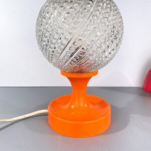 May include: A vintage table lamp with a clear glass globe shade with a textured pattern and an orange plastic base.