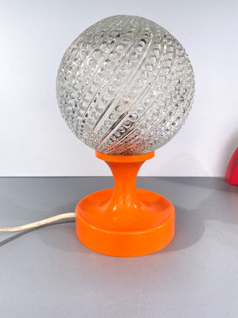 May include: A vintage table lamp with a clear glass globe shade with a textured pattern and an orange plastic base.