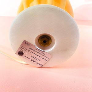 May include: A white ceramic lamp base with a gold screw and a label that reads "GEORGIA JACOB, Mod&egrave;le d&eacute;pos&eacute; ATH&Eacute;NA Puis, max. 40 W tube x Made in France Canforme norme europ&eacute;enne EN 60598 220 V". The lamp base is topped with a yellow glass lampshade shaped like a flame.