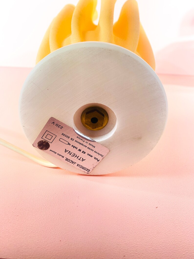 May include: A white ceramic lamp base with a gold screw and a label that reads "GEORGIA JACOB, Mod&egrave;le d&eacute;pos&eacute; ATH&Eacute;NA Puis, max. 40 W tube x Made in France Canforme norme europ&eacute;enne EN 60598 220 V". The lamp base is topped with a yellow glass lampshade shaped like a flame.