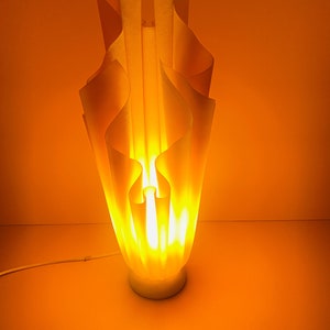 May include: A white, wavy, sculptural lamp with a warm, yellow glow. The lamp is lit and casts a warm light on the orange background.