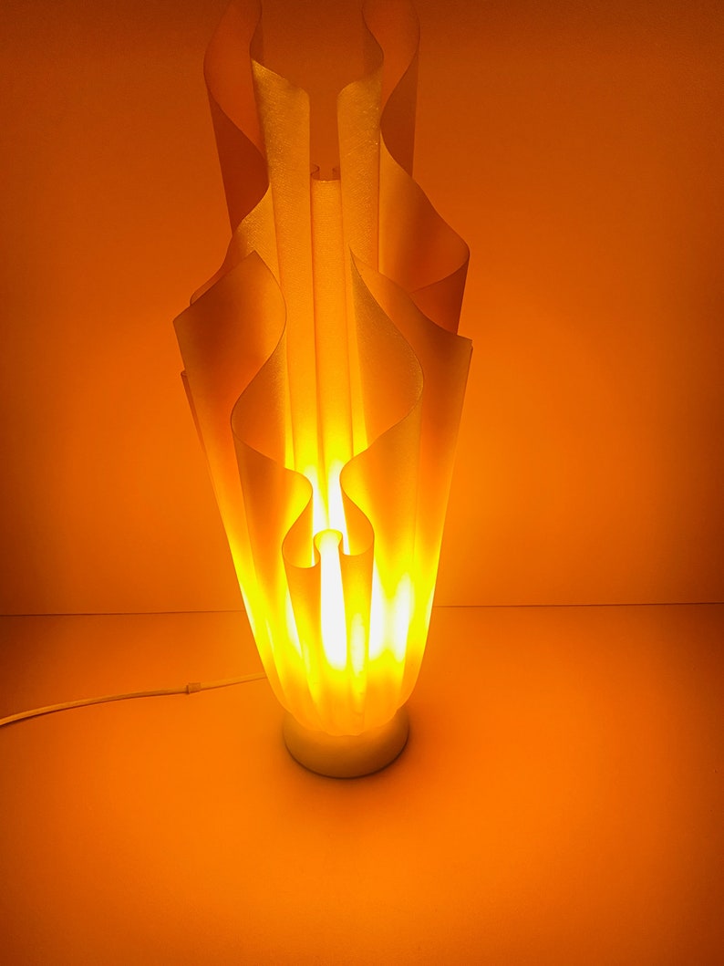 May include: A white, wavy, sculptural lamp with a warm, yellow glow. The lamp is lit and casts a warm light on the orange background.
