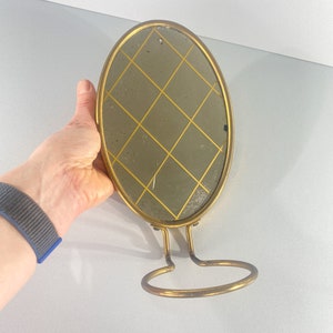 Puede incluir: Un espejo ovalado dorado con un patrón de diamante en el vidrio. El espejo está unido a un soporte de metal dorado con un anillo en la parte inferior.