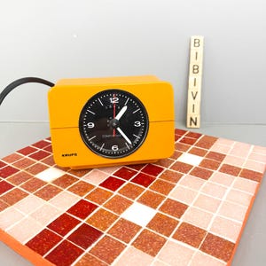 May include: A vintage orange KRUPS Comfortime alarm clock with a black clock face and white hands. The clock sits on a mosaic tile surface with red, orange, and white squares. A black power cord extends from the clock.