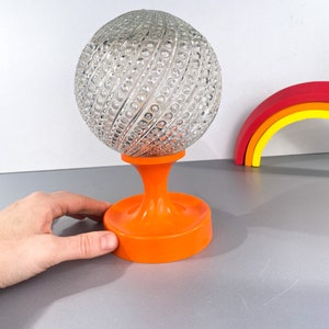 May include: A clear glass globe lamp with a textured surface sits on an orange plastic base. The globe has a swirling pattern of raised dots.