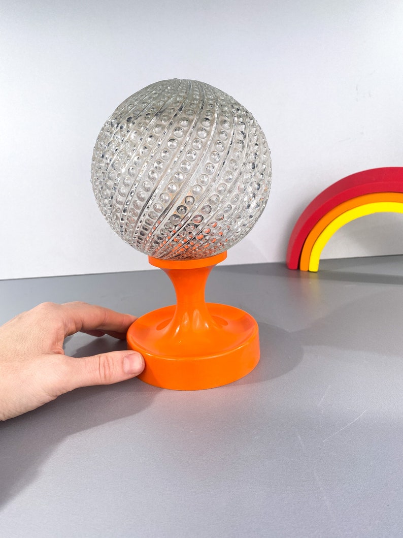 May include: A clear glass globe lamp with a textured surface sits on an orange plastic base. The globe has a swirling pattern of raised dots.