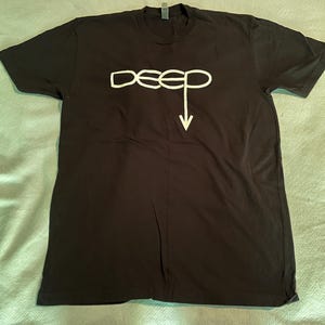 May include: A black short-sleeve t-shirt featuring the word deep printed in white across the chest. The letter p in the word deep extends downward into a white arrow pointing toward the bottom hem of the garment. The shirt has a crew neckline.