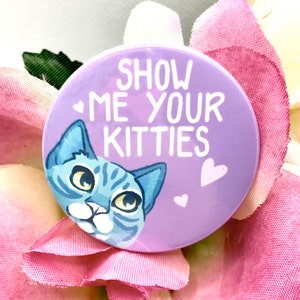 Puede incluir: Botón morado con el texto "Show me your kitties" en blanco. Un gato azul atigrado de dibujos animados con ojos amarillos asoma por el lado izquierdo del botón. Hay tres corazones rosas en el botón.