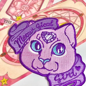 Könnte beinhalten: Lilafarbener gestickter Aufnäher mit einer Cartoon-Katze mit einem großen Diamanten auf der Stirn. Die Katze trägt einen lilafarbenen Schal mit den gestickten Worten "Meow Great" und "Girl".