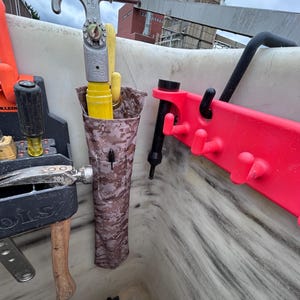 Pu&ograve; includere: Un contenitore per riporre gli attrezzi con vari strumenti. In alto, un gancio in metallo argentato, con un manico giallo e una custodia in tessuto mimetico. Un portautensili in plastica rossa a destra. Altri strumenti sono visibili.