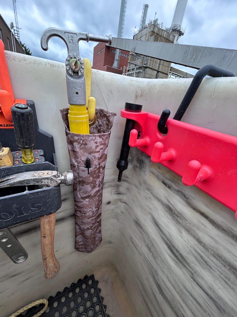 Pu&ograve; includere: Un contenitore per riporre gli attrezzi con vari strumenti. In alto, un gancio in metallo argentato, con un manico giallo e una custodia in tessuto mimetico. Un portautensili in plastica rossa a destra. Altri strumenti sono visibili.