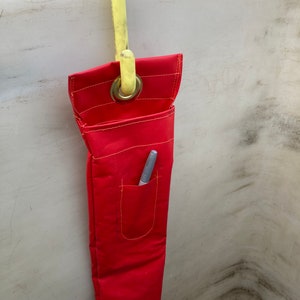 May include: A red tool bag with a yellow hook hanging from a white surface. The bag has a pocket with a silver pen inside.