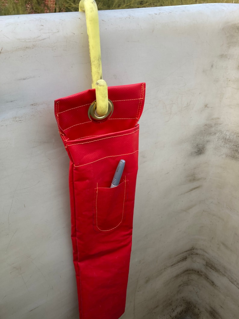 May include: A red tool bag with a yellow hook hanging from a white surface. The bag has a pocket with a silver pen inside.