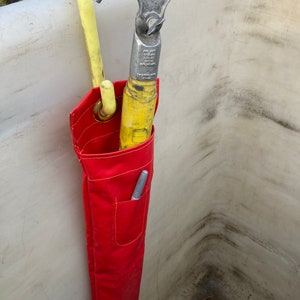 May include: A red tool bag with a yellow hook and a silver hook hanging from it. The bag is hanging on a white surface.