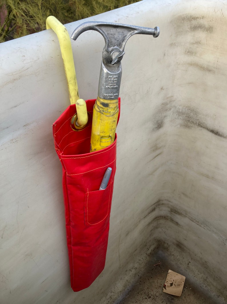 May include: A red tool bag with a yellow hook and a silver hook hanging from it. The bag is hanging on a white surface.