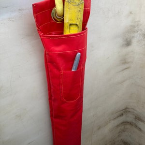 May include: A red canvas tool bag with a yellow and silver tool inside. The bag has a small pocket on the front with a pen inside.