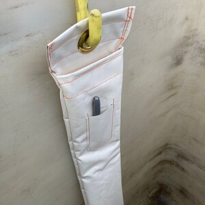 K&ouml;nnte beinhalten: Wei&szlig;er Werkzeughalter aus Stoff mit orangefarbenen N&auml;hten und einer kleinen Tasche, der an einem gelben Haken h&auml;ngt. Ein grauer Stift befindet sich in der Tasche. Der Halter besteht aus strapazierf&auml;higem, strukturiertem Material.