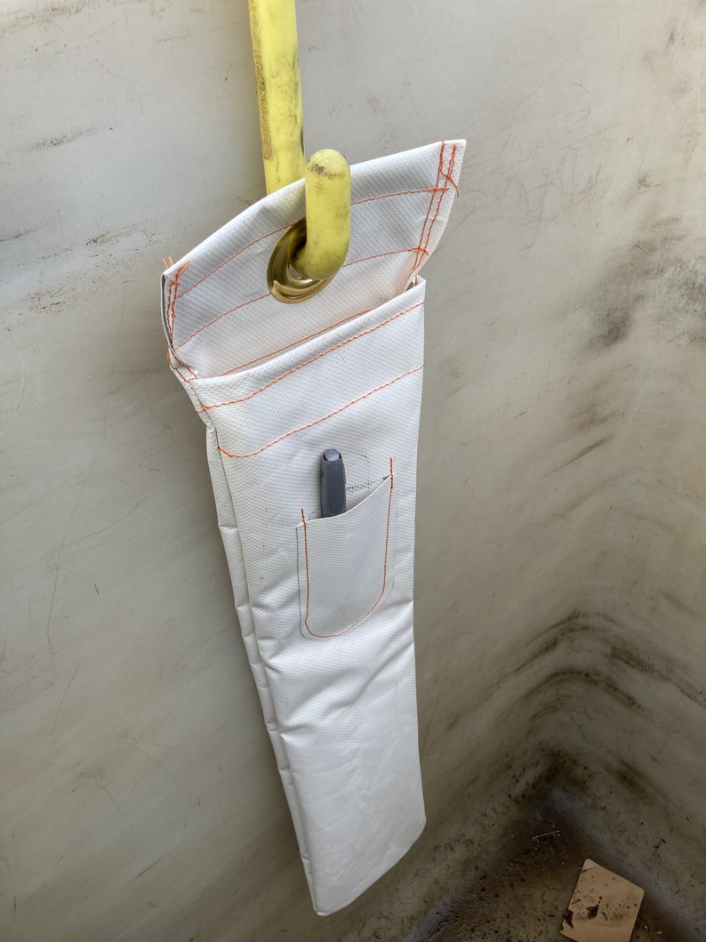 K&ouml;nnte beinhalten: Wei&szlig;er Werkzeughalter aus Stoff mit orangefarbenen N&auml;hten und einer kleinen Tasche, der an einem gelben Haken h&auml;ngt. Ein grauer Stift befindet sich in der Tasche. Der Halter besteht aus strapazierf&auml;higem, strukturiertem Material.