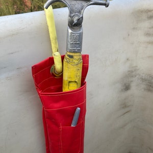 May include: A red canvas tool bag with a yellow and silver hook tool inside. The bag has a small pocket with a silver pen inside. The text on the tool reads "Do Not Leave Hook Unsecured".