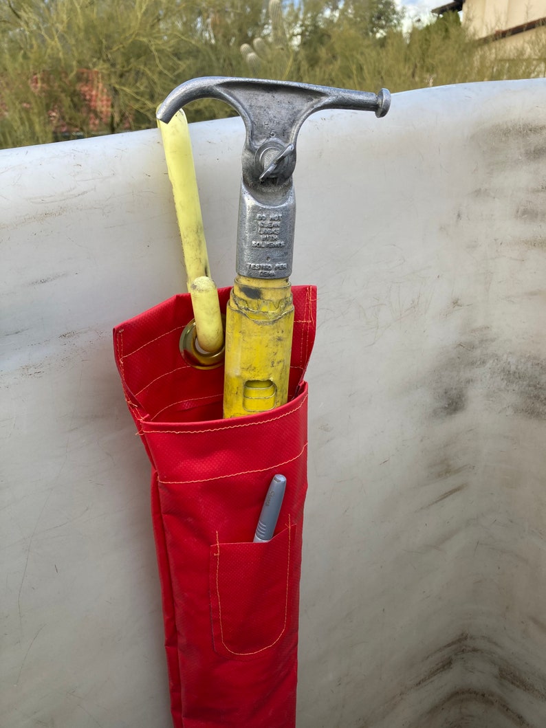May include: A red canvas tool bag with a yellow and silver hook tool inside. The bag has a small pocket with a silver pen inside. The text on the tool reads "Do Not Leave Hook Unsecured".