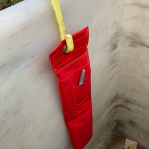 May include: A red tool bag with a pocket and a yellow hook hanging from a white surface.