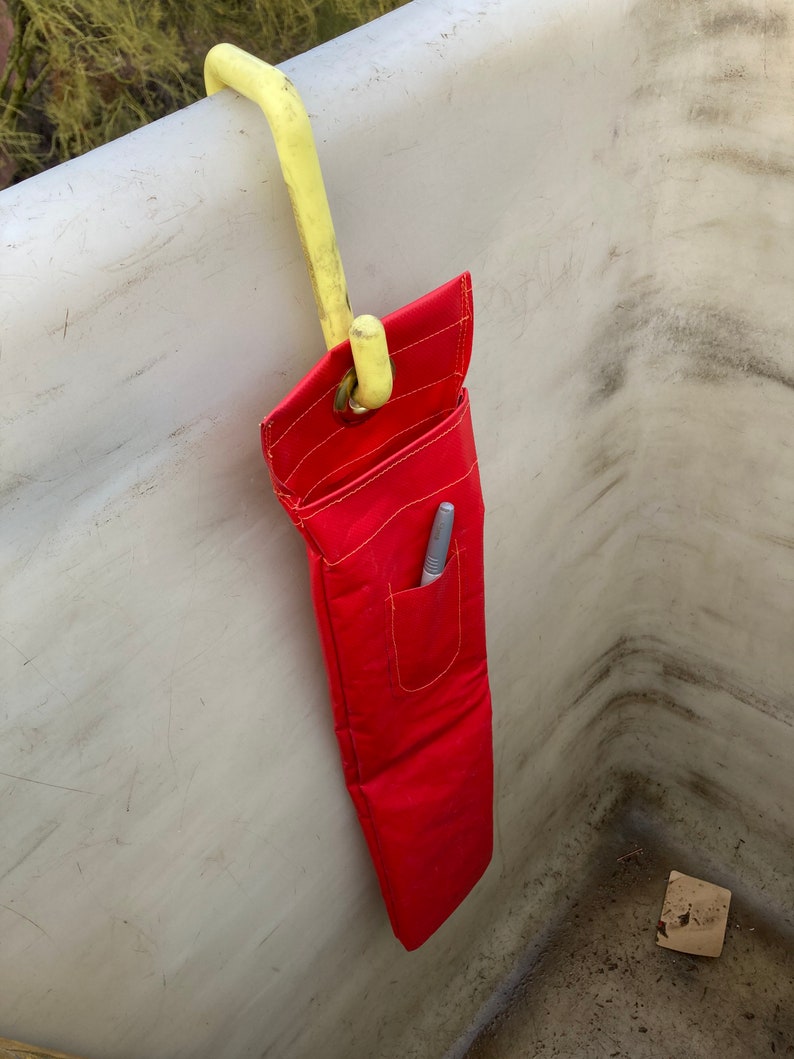 May include: A red tool bag with a pocket and a yellow hook hanging from a white surface.