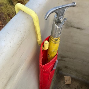 May include: A red tool bag with a yellow and silver hook tool hanging inside. The bag is hanging on a yellow hook attached to a white surface.