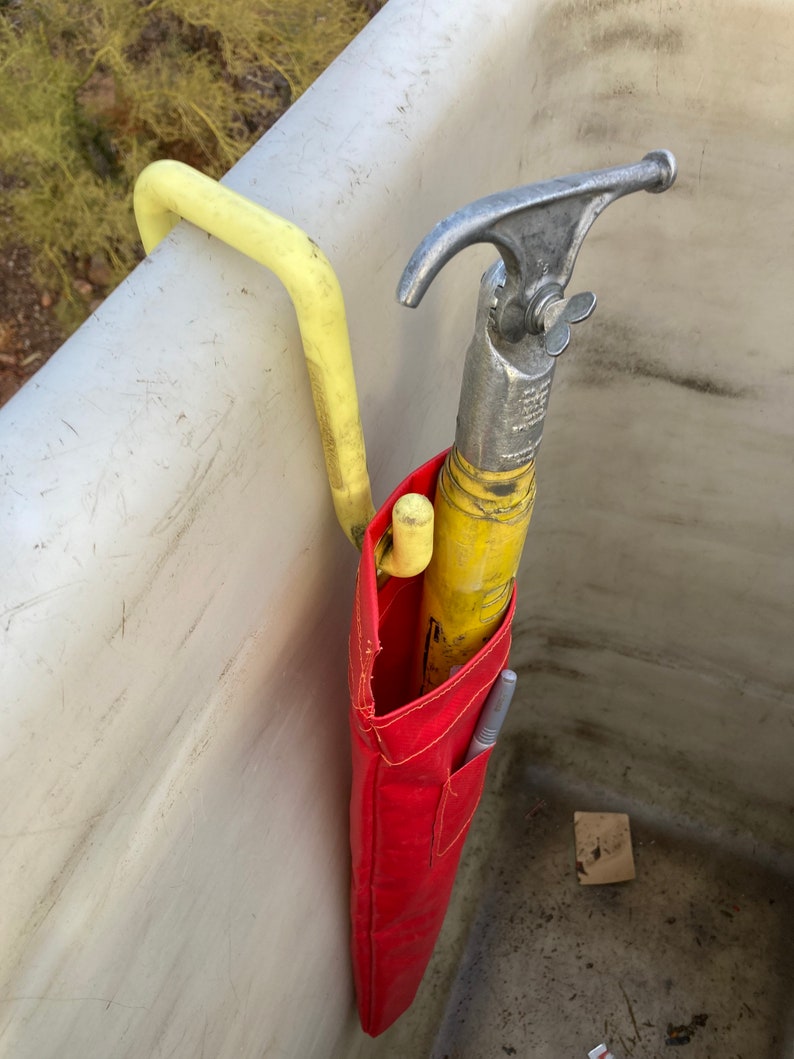 May include: A red tool bag with a yellow and silver hook tool hanging inside. The bag is hanging on a yellow hook attached to a white surface.