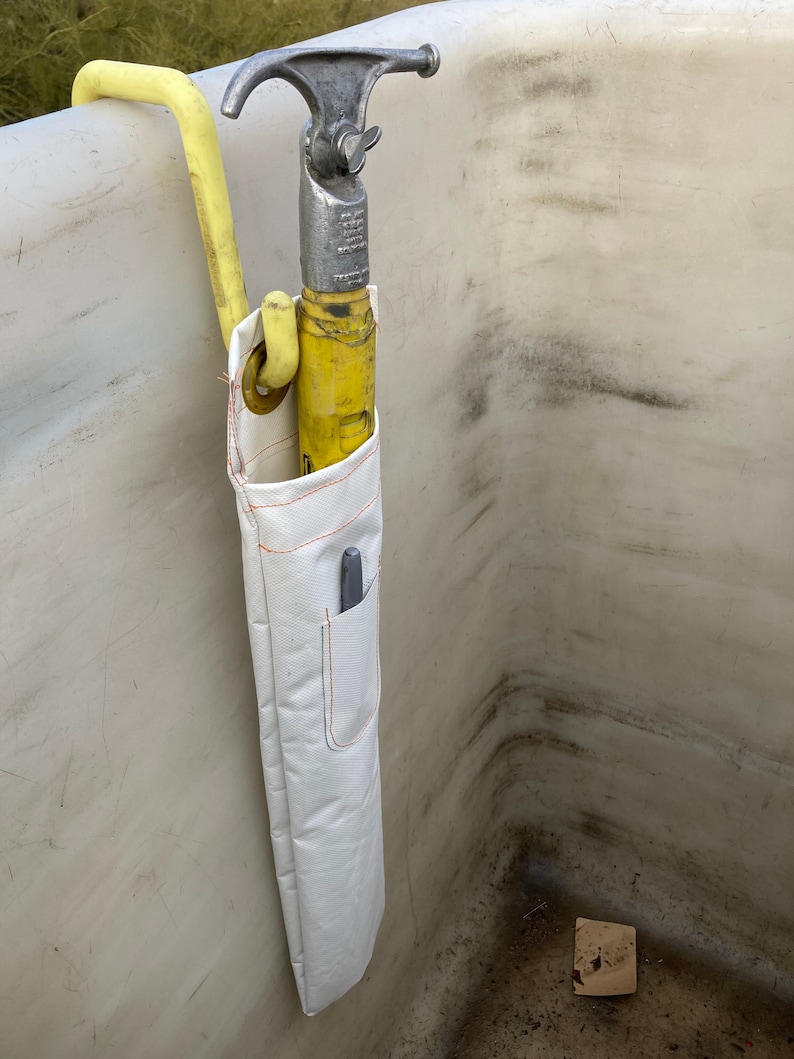 K&ouml;nnte beinhalten: Ein gelbes und silbernes Feuerwehrwerkzeug mit einer wei&szlig;en Segeltuchscheide. Das Werkzeug hat einen Haken und ein spitzes Ende. Die Scheide hat eine Tasche mit einem Stift darin. Das Werkzeug h&auml;ngt an einer wei&szlig;en Oberfl&auml;che.