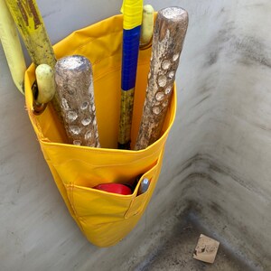 May include: A yellow canvas tool bag with multiple pockets, hanging on a wall. The bag is filled with various tools, including a yellow wooden handled tool with blue tape, and three wooden handled tools with brown and black markings.