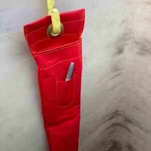 May include: A red fabric tool bag with a yellow hook and a small pocket with a pen inside. The bag is hanging on a white wall.