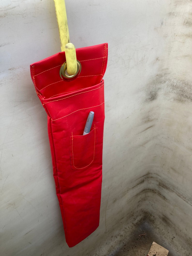 May include: A red fabric tool bag with a yellow hook and a small pocket with a pen inside. The bag is hanging on a white wall.