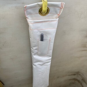 K&ouml;nnte beinhalten: Werkzeughalter aus wei&szlig;em Stoff mit orangefarbenen N&auml;hten und einer kleinen Tasche. Ein grauer Stift befindet sich in der Tasche. Der Halter wird von einer gelben Stange durch eine Messing&ouml;se gehalten. Der Werkzeughalter ist f&uuml;r Werkzeuge konzipiert.