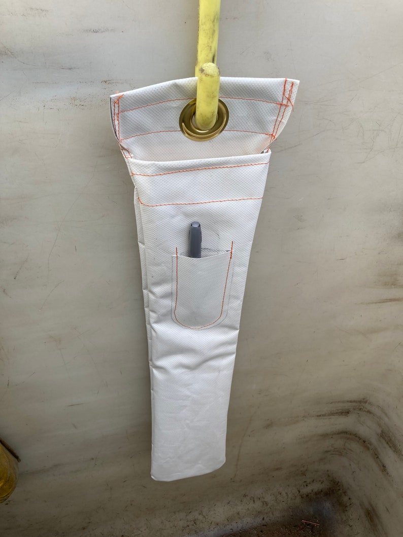 K&ouml;nnte beinhalten: Werkzeughalter aus wei&szlig;em Stoff mit orangefarbenen N&auml;hten und einer kleinen Tasche. Ein grauer Stift befindet sich in der Tasche. Der Halter wird von einer gelben Stange durch eine Messing&ouml;se gehalten. Der Werkzeughalter ist f&uuml;r Werkzeuge konzipiert.