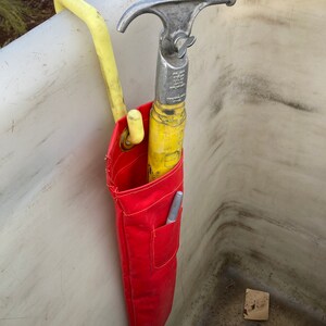 May include: A red canvas tool bag with a yellow and silver hook tool hanging inside. The bag is attached to a white hook.