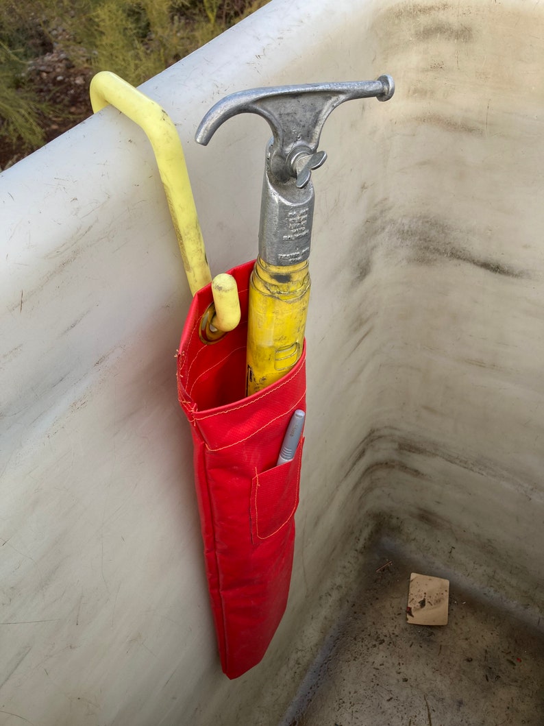 May include: A red canvas tool bag with a yellow and silver hook tool hanging inside. The bag is attached to a white hook.