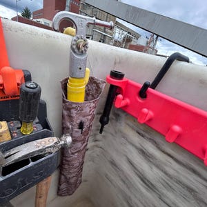Pu&ograve; includere: Una collezione di strumenti, tra cui un martello, un cacciavite e una chiave inglese, &egrave; visibile in un'area di lavoro. Un porta attrezzi mimetico contiene un tubo giallo e una penna. &Egrave; presente anche un supporto in plastica rossa con ganci.