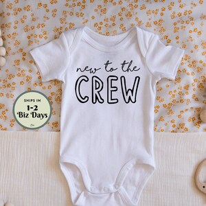 May include: White baby onesie with the text "new to the CREW" in black. The onesie is laid on a cream-colored surface with a floral pattern. A wooden rattle and a wooden elephant-shaped teether are also present.