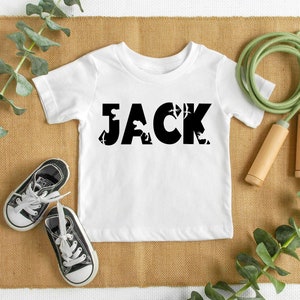 May include: A white short-sleeved t-shirt featuring the name JACK printed in large black letters. The letters contain silhouettes of various dinosaurs. The shirt is displayed alongside a pair of black and white sneakers and a jump rope.